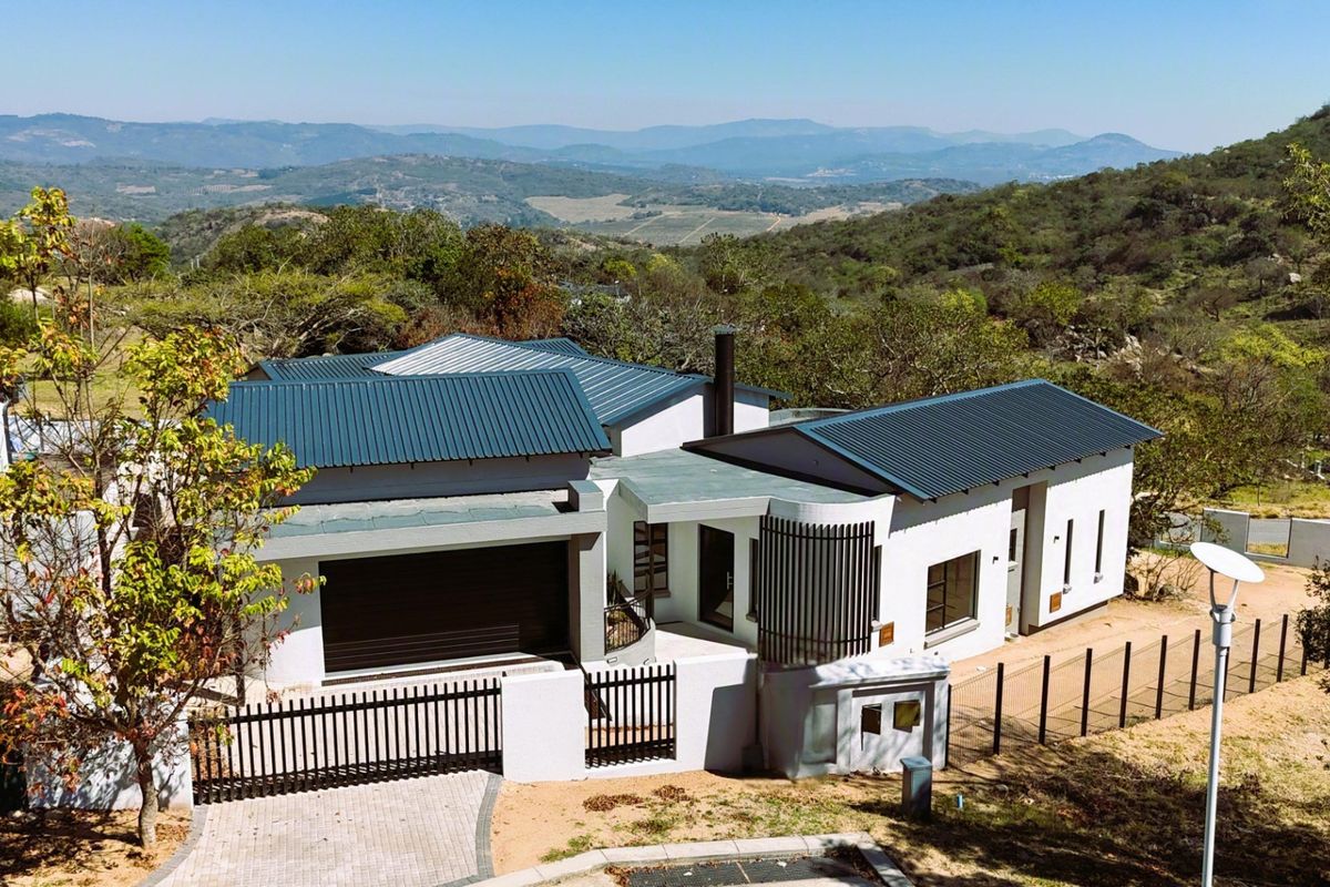 Aerial view of a modern residence featuring a corrugated metal roof, double garage, and paved driveway, set against a scenic mountainous landscape.