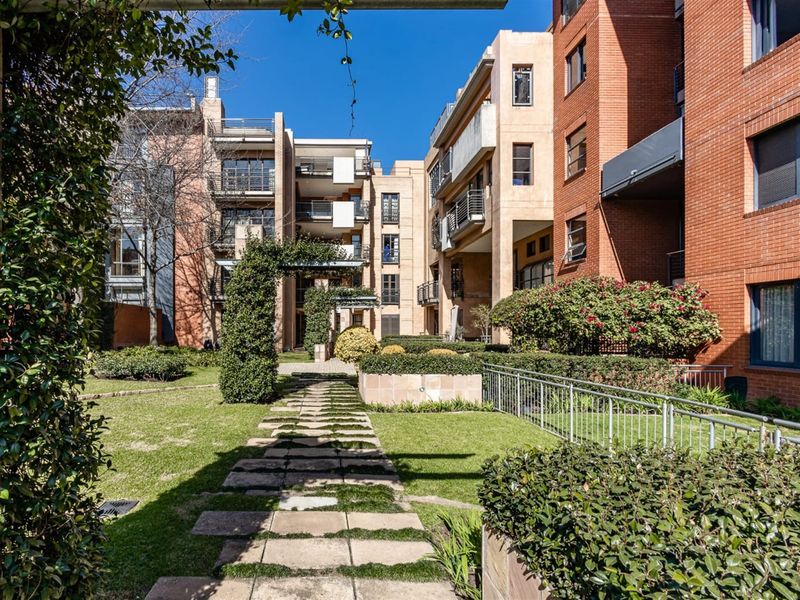 A well-maintained communal garden area featuring a stone pathway, manicured hedging, and brick apartment building facades with metal balcony railings.