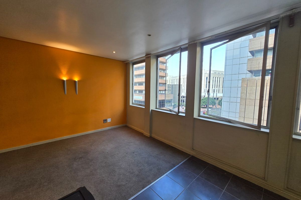 An empty living area featuring large windows with city views, wall-mounted lighting, and a combination of carpeted and tiled flooring.