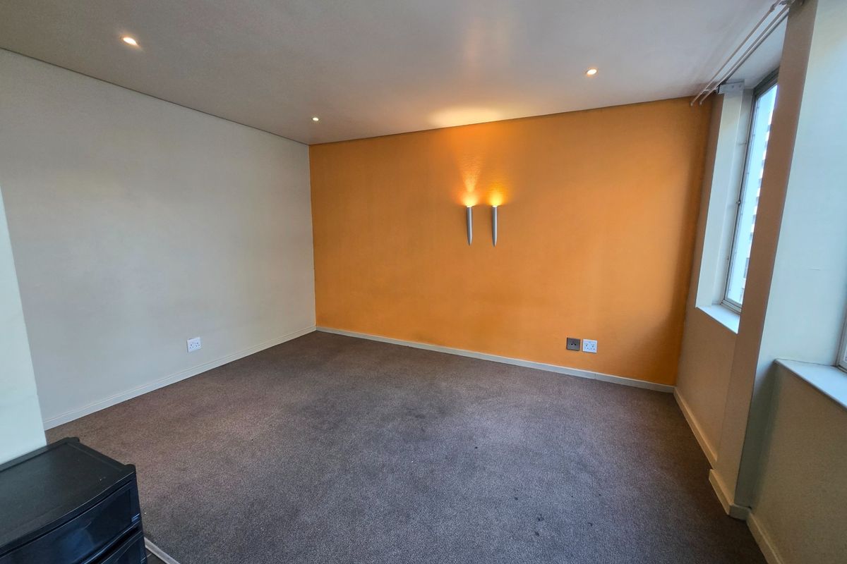A spacious, empty bedroom featuring neutral and accent-colored walls, recessed ceiling lighting, wall-mounted sconces, and carpeted flooring.