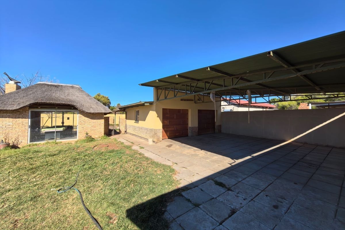 Rear exterior view featuring a double carport with metal roofing, paved driveway area, and a separate brick structure with a thatched roof.
