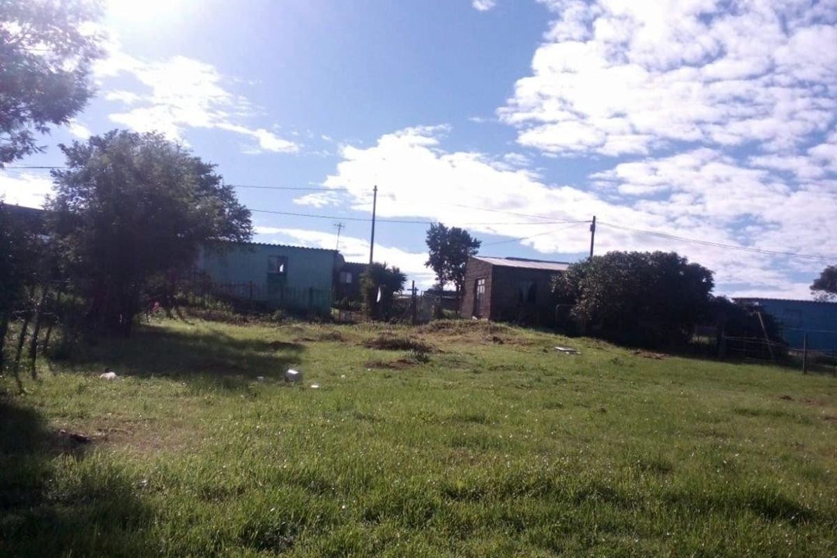 A view of a vacant residential plot featuring grassy terrain with surrounding structures visible in the background.