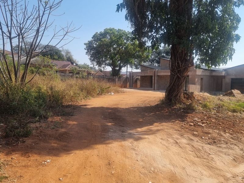 A view of a residential plot featuring an unfinished building structure, unpaved access path, and mature trees on the property.