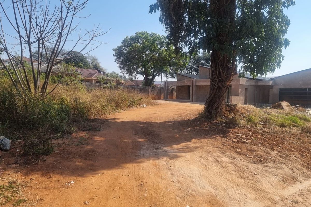 A view of a residential plot featuring an unfinished building structure, unpaved access path, and mature trees on the property.