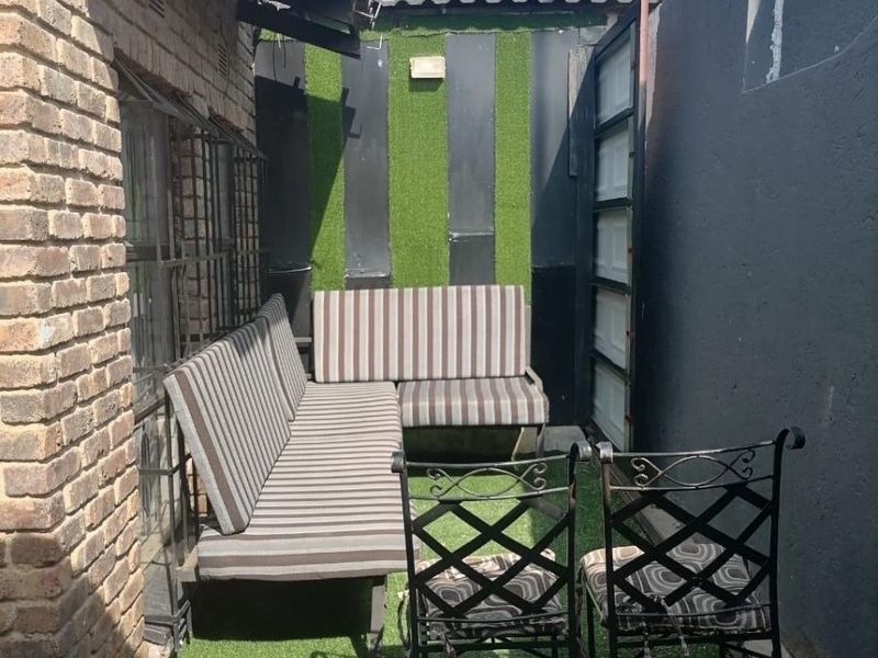 A compact, paved outdoor patio area featuring artificial grass, brick walling, and a covered section, suitable for small-scale outdoor seating.
