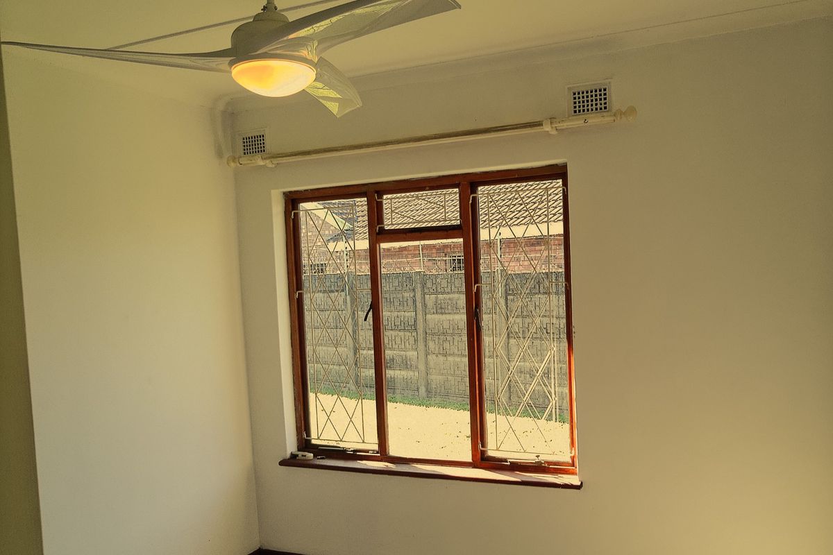 A bright bedroom featuring a ceiling fan, tiled flooring, and a large window with security bars overlooking the rear yard.