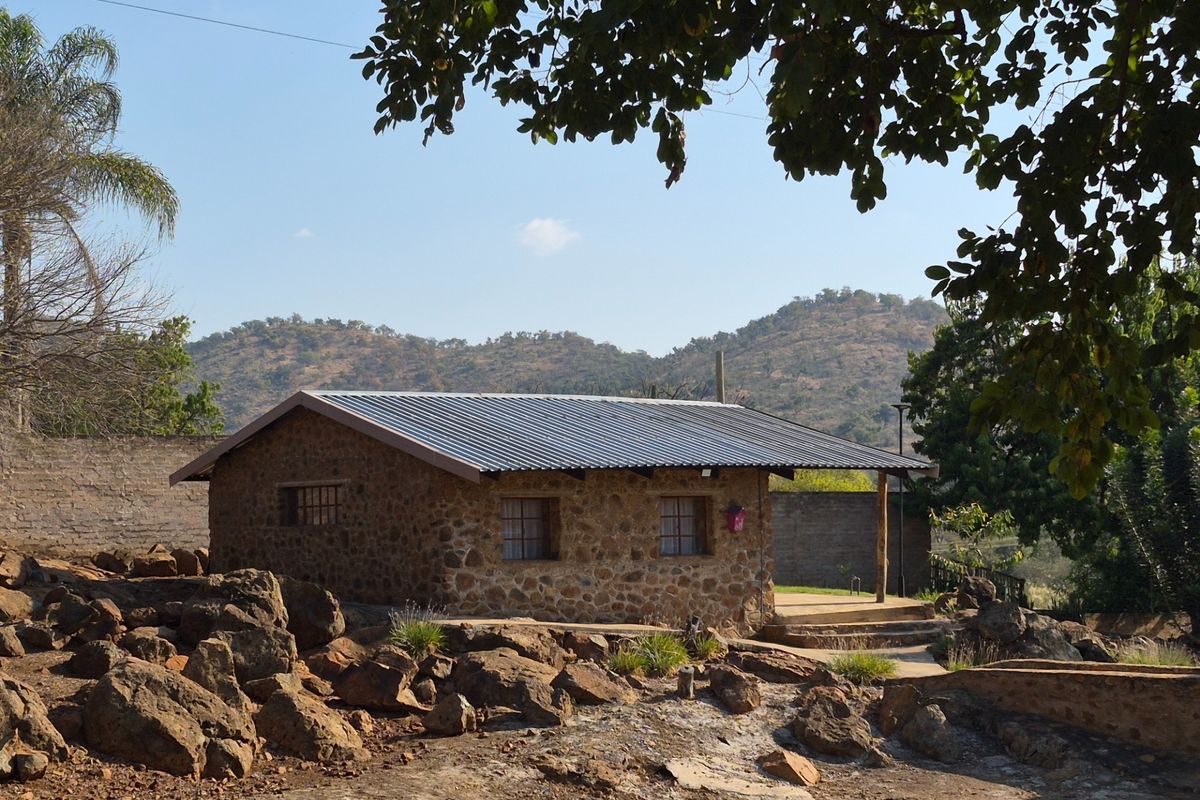 A rustic stone-clad cottage featuring a corrugated metal roof, set within a natural rocky landscape with mountain views.