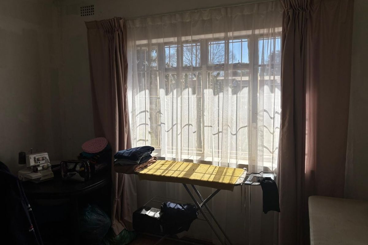 A bedroom featuring a large window with security bars, neutral curtains, and basic wall finishes, offering potential for renovation and modernization.