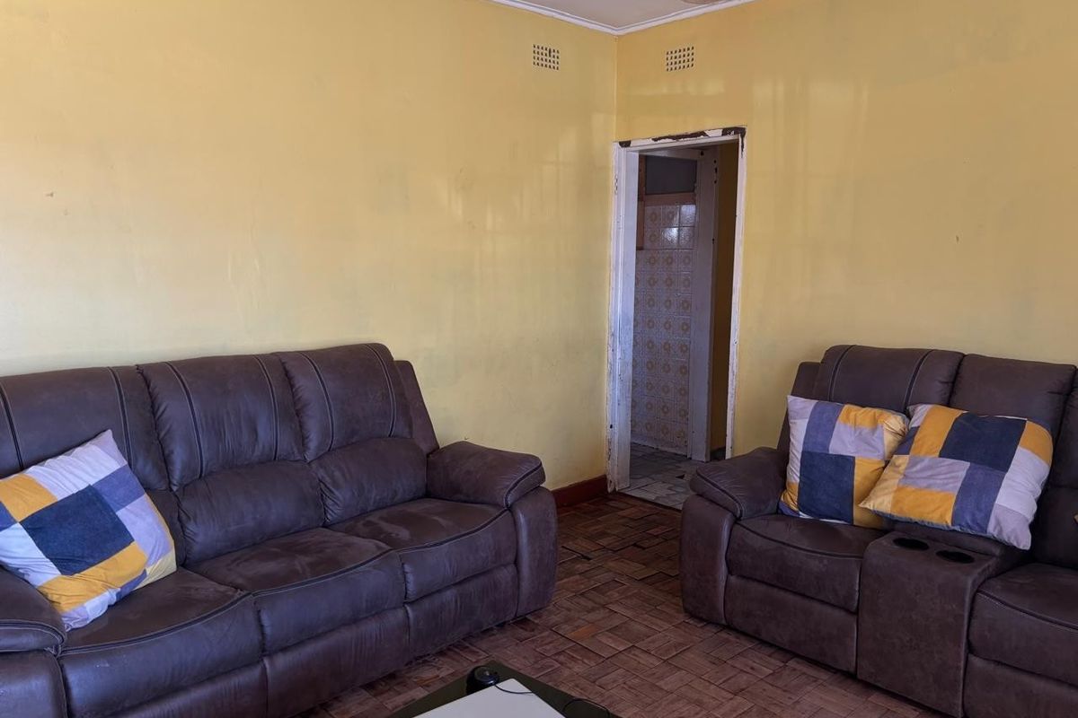 A living room featuring parquet flooring and yellow walls, with visible signs of wear and ceiling water damage, leading into an adjacent tiled room.