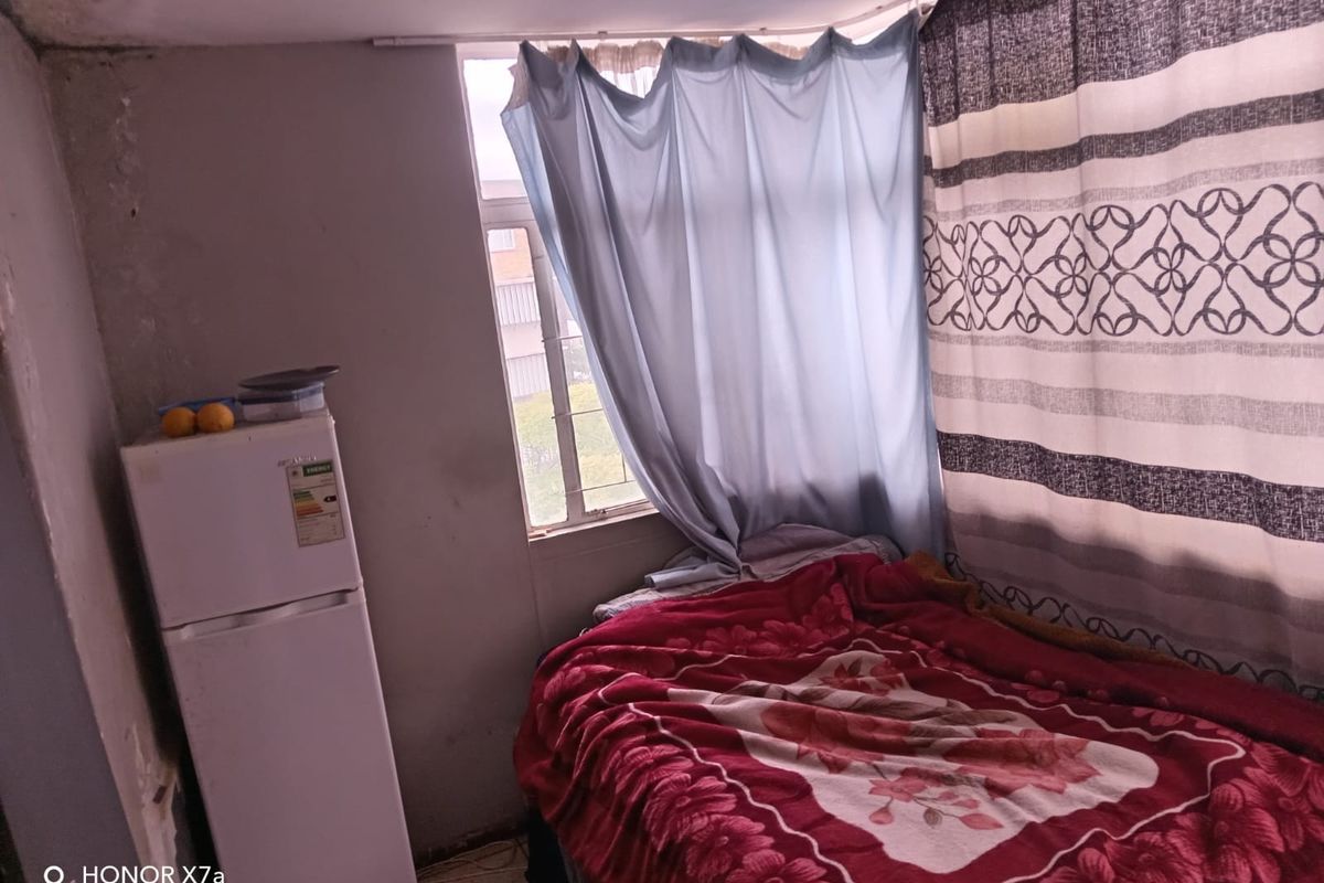 A compact bedroom featuring a window with curtains and space for a bed and refrigerator.