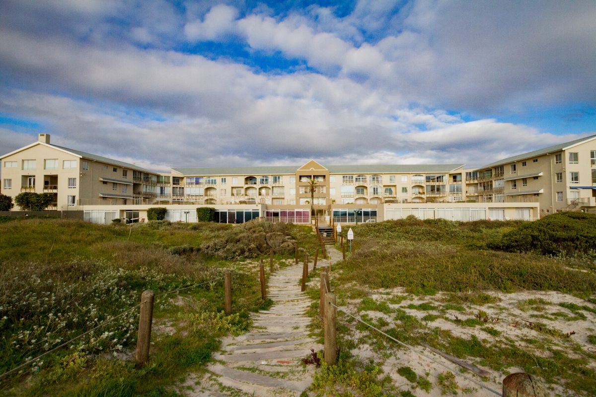 A view of the apartment complex exterior from the beach path, featuring multi-story residential units with balconies and landscaped coastal dunes.