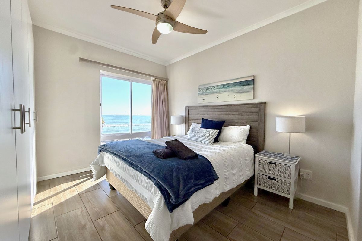 A bright bedroom featuring wood-look tile flooring, a ceiling fan, built-in wardrobes, and a large window offering scenic ocean views.