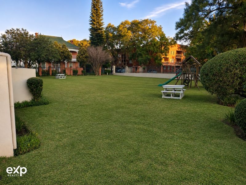 A well-maintained communal garden area featuring a large lawn, mature trees, and a children's play structure within the apartment complex grounds.