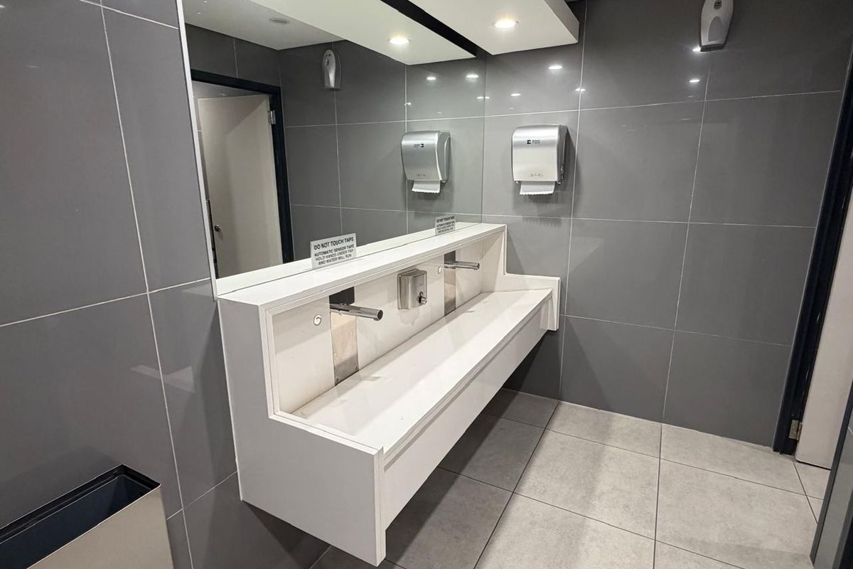 Modern office restroom featuring a sleek, wall-mounted trough sink with sensor-operated taps, large format grey wall tiles, and integrated mirror lighting.