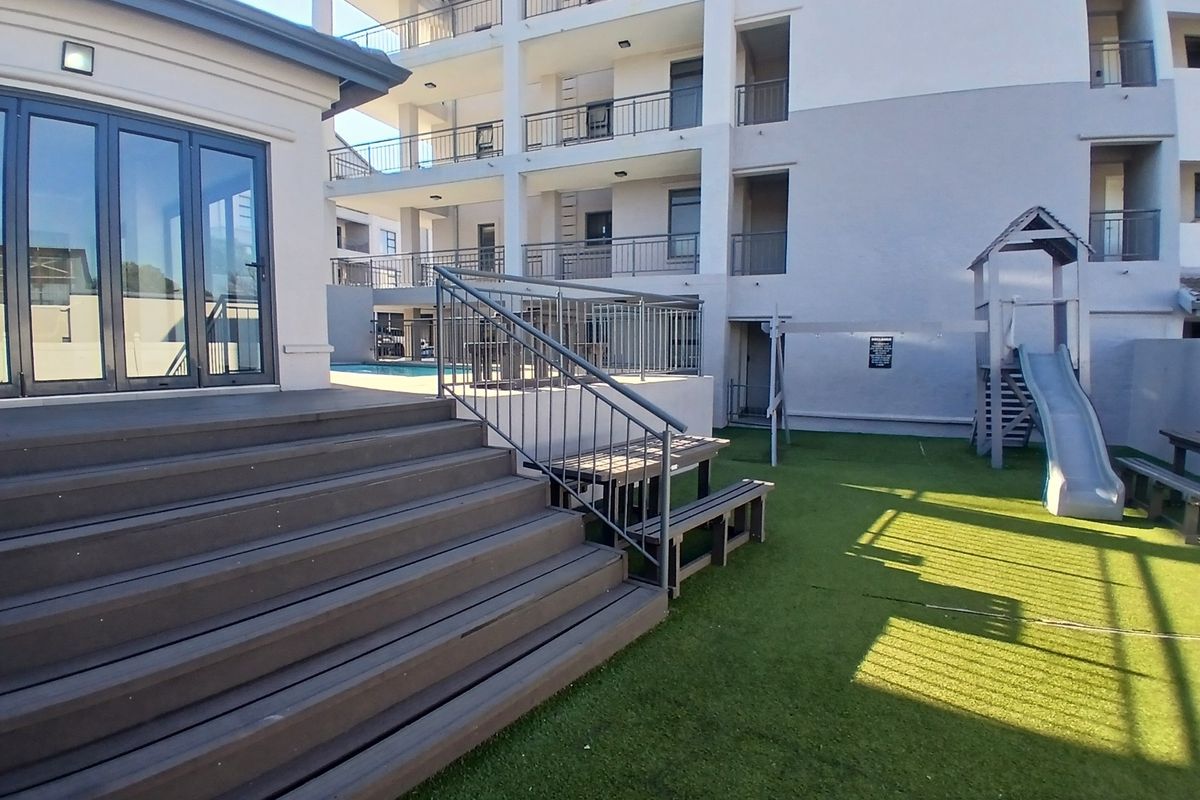 Communal outdoor area featuring a raised timber deck, synthetic lawn, and a children's play structure set against the apartment building facade.