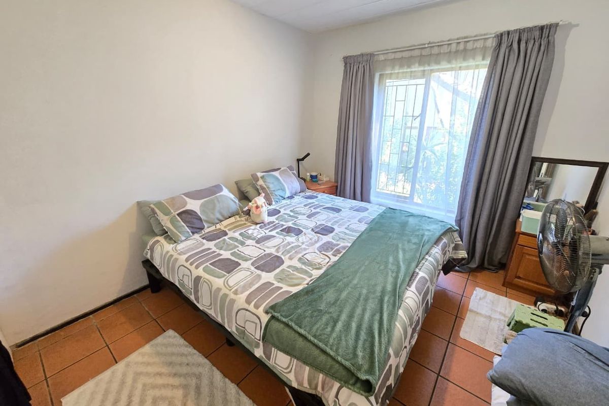 A functional bedroom featuring terracotta floor tiles, a large window with security bars, and neutral wall finishes.