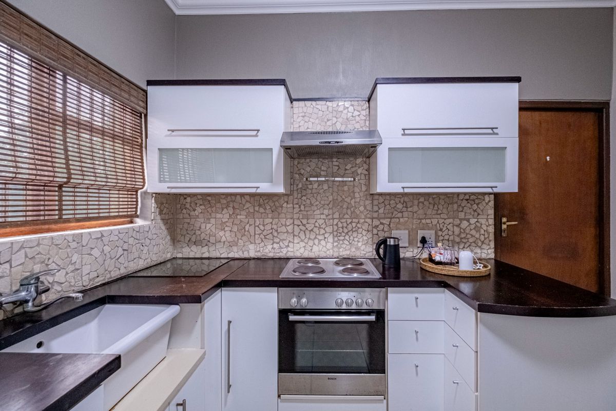 A compact, well-equipped kitchen featuring white cabinetry, dark countertops, a built-in oven, electric hob, extractor fan, and a classic farmhouse-style sink.