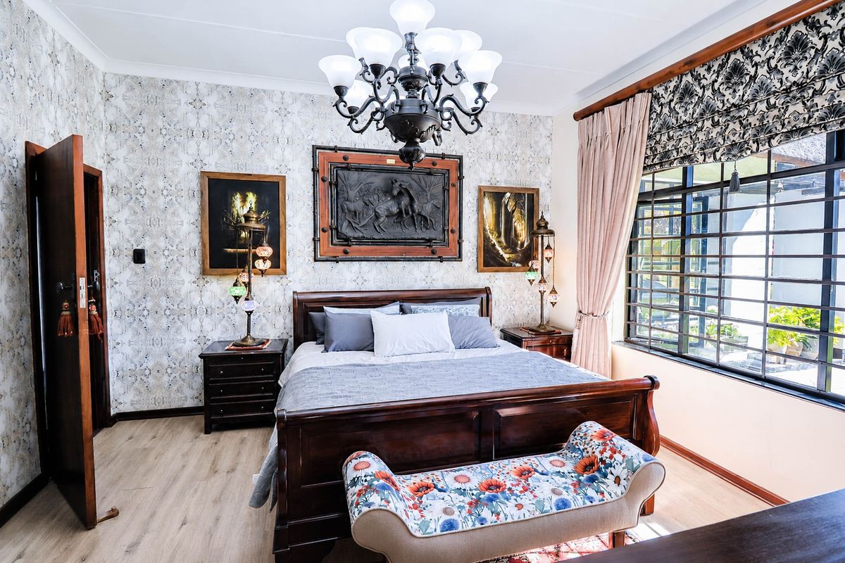 A well-maintained bedroom featuring laminate flooring, decorative wallpaper, a central chandelier, and large windows fitted with security bars.