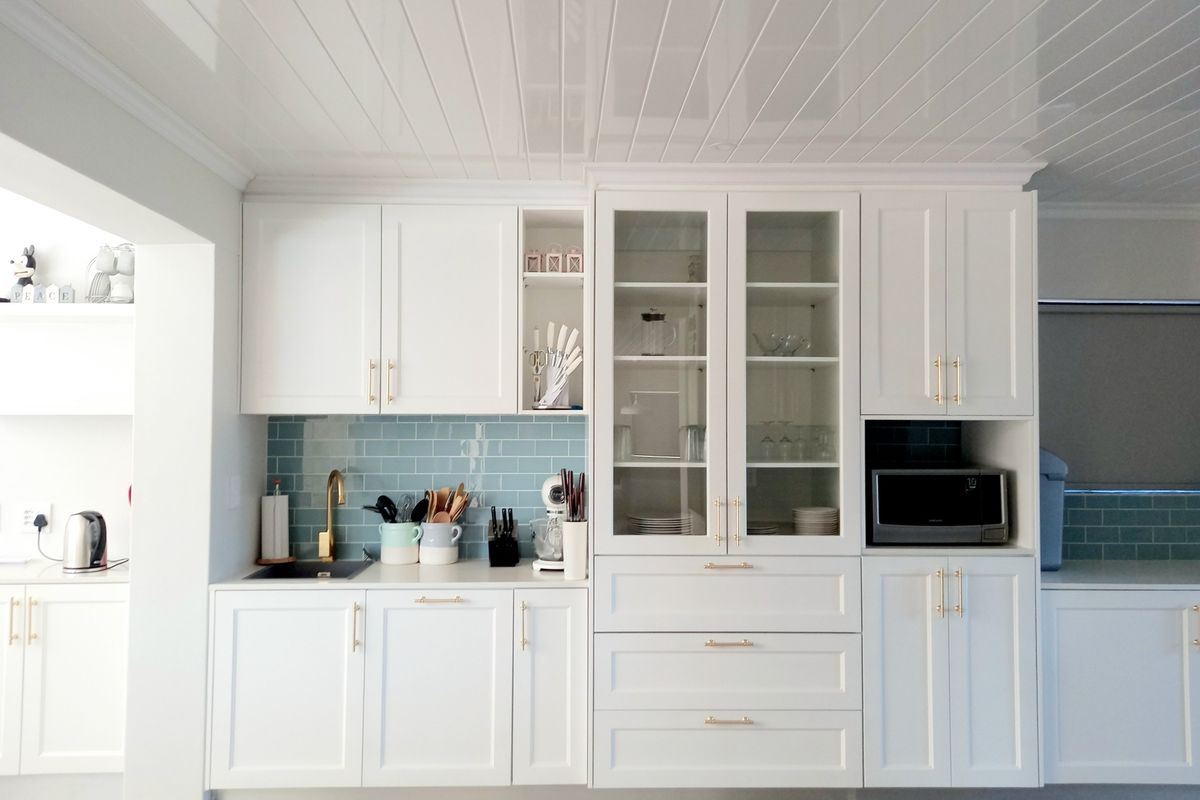 A bright, modern kitchen featuring white cabinetry, glass-front display cupboards, a blue subway tile backsplash, and gold hardware accents.