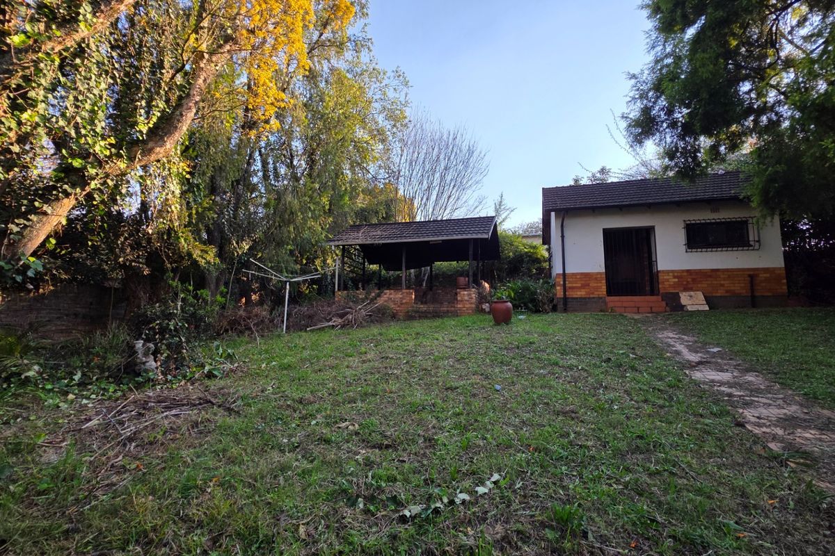 A rear garden view featuring a small outbuilding with a covered patio, brick braai area, and a paved pathway leading to the structure.