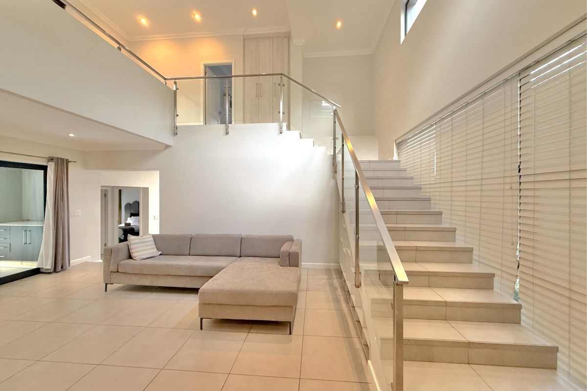 Spacious open-plan living area featuring modern tiled flooring, a striking glass staircase balustrade, and abundant natural light through large windows.