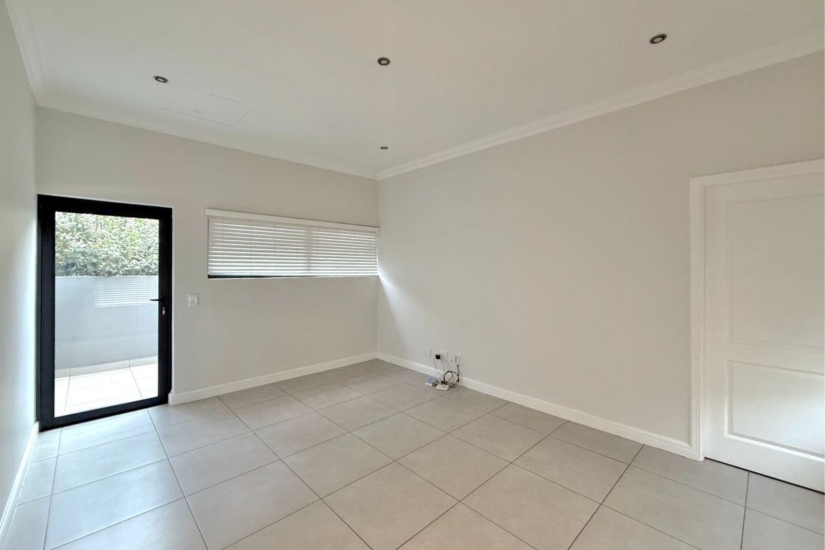 A bright, empty bedroom featuring neutral tiled flooring, recessed downlighting, and a glass door providing direct access to an outdoor area.