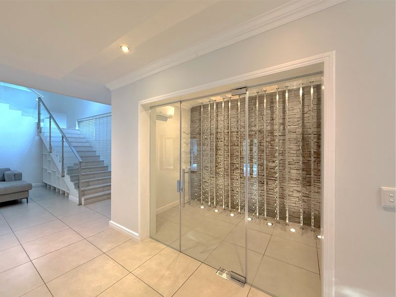 A modern, climate-controlled glass-enclosed wine cellar featuring a stone accent wall, custom metal racking, and recessed lighting, adjacent to a contemporary staircase.