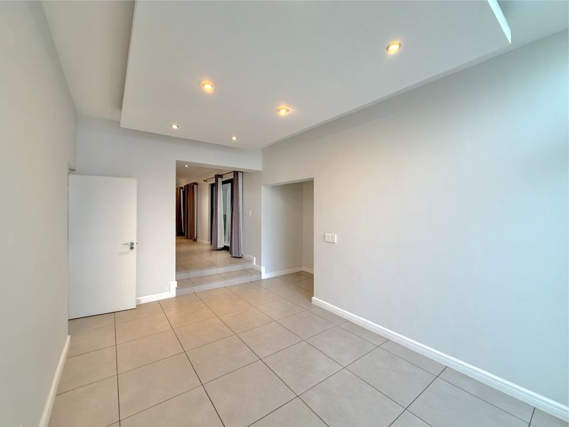 A bright, empty living area featuring neutral tiled flooring, a recessed ceiling with integrated downlights, and a step leading to an adjacent space.