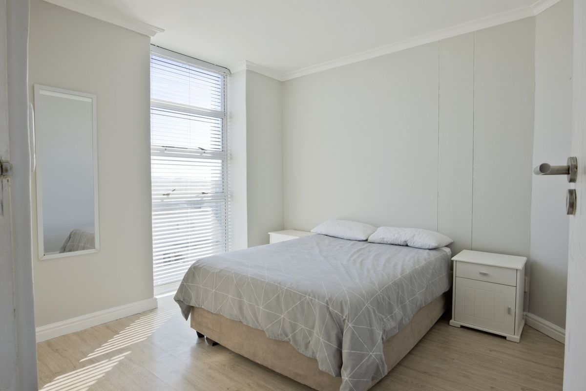 A bright and neutral bedroom featuring light wood-look flooring, crown molding, and a large window with fitted blinds.