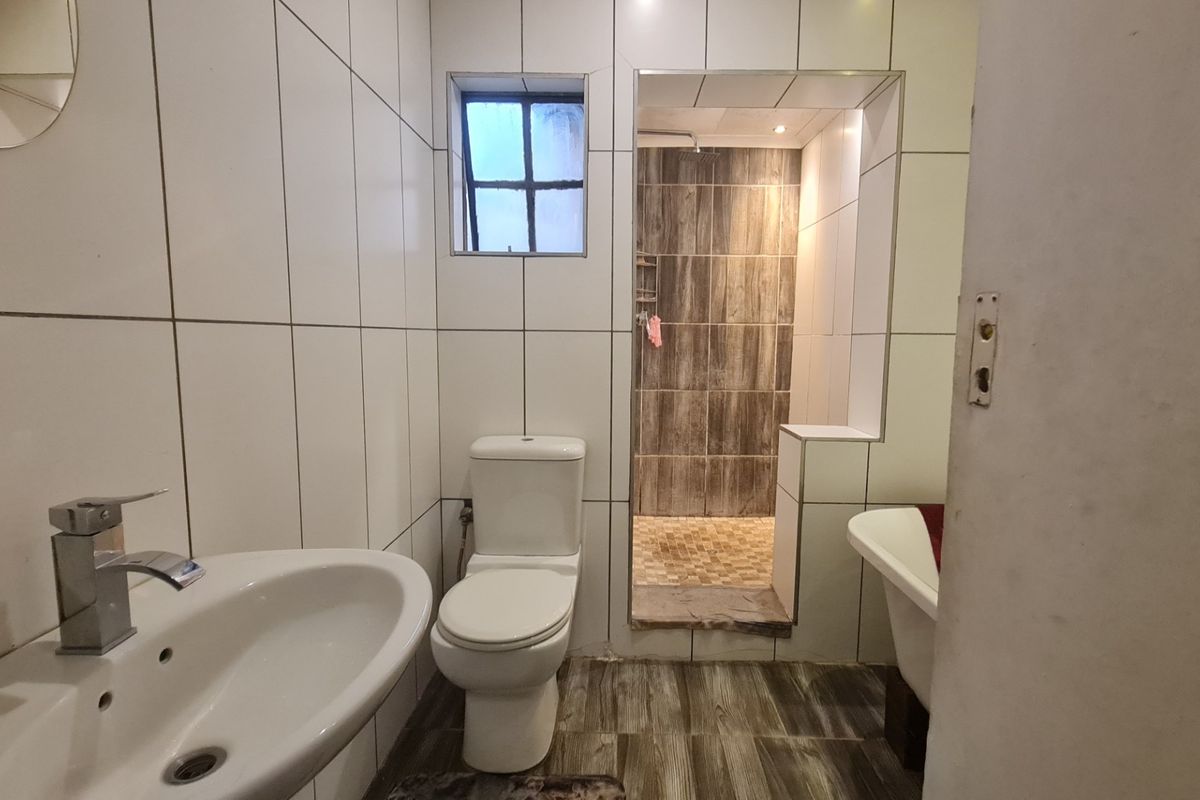 A functional bathroom featuring a walk-in shower with wood-look tiling, a pedestal basin, and a toilet, finished with white wall tiles.