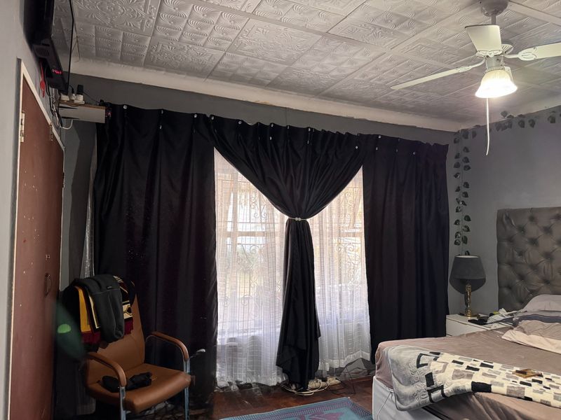 A bedroom featuring decorative ceiling tiles, a ceiling fan, and security bars on the window, with visible wear on the wall finishes.