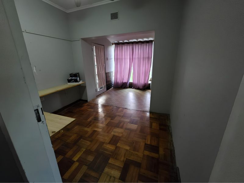 An empty living area featuring original parquet flooring, built-in shelving, and a transition space leading to a windowed area with curtains.