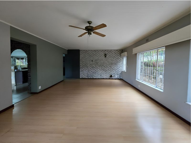 Spacious, empty living room featuring a brick accent wall, ceiling fan, and large windows with security bars, offering a versatile blank canvas.