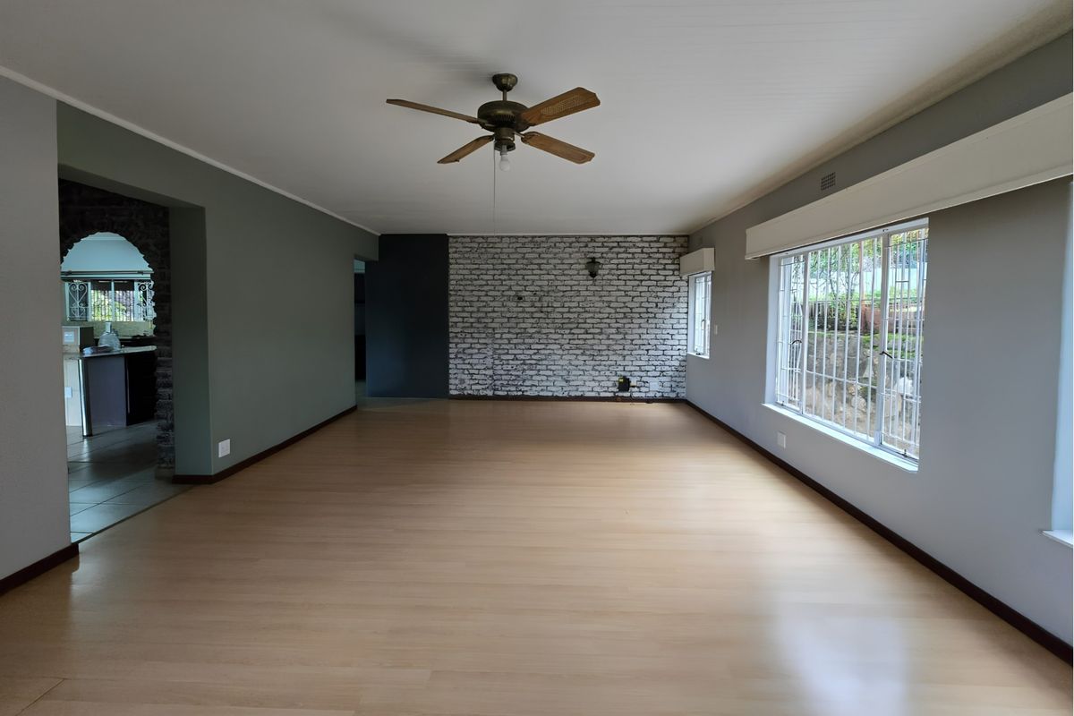 Spacious, empty living room featuring a brick accent wall, ceiling fan, and large windows with security bars, offering a versatile blank canvas.
