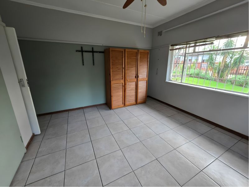 A spacious, empty bedroom featuring neutral tiled flooring, a ceiling fan, and a built-in wooden cupboard with security bars on the window.