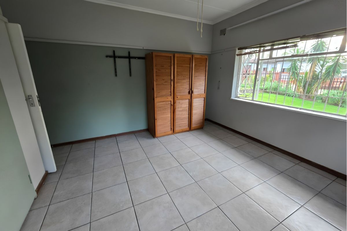 A spacious, empty bedroom featuring neutral tiled flooring, a ceiling fan, and a built-in wooden cupboard with security bars on the window.