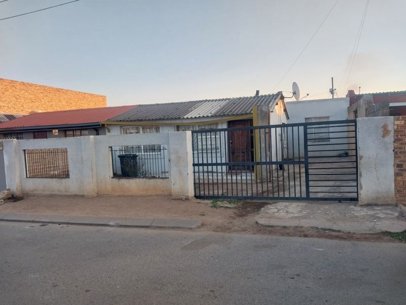 Front exterior view of a single-story house featuring a gated entrance, perimeter wall, and corrugated roof sheeting.