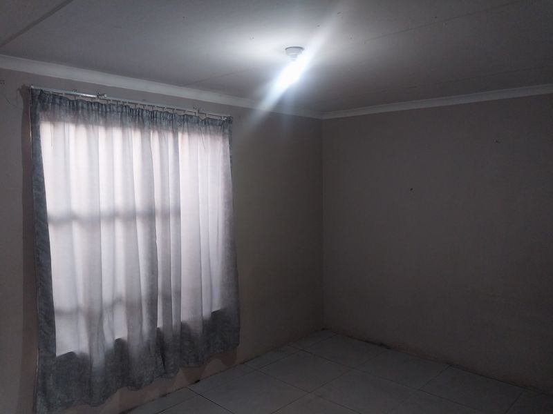 A simple, empty bedroom featuring tiled flooring, neutral walls, and a window with fitted curtain rails.