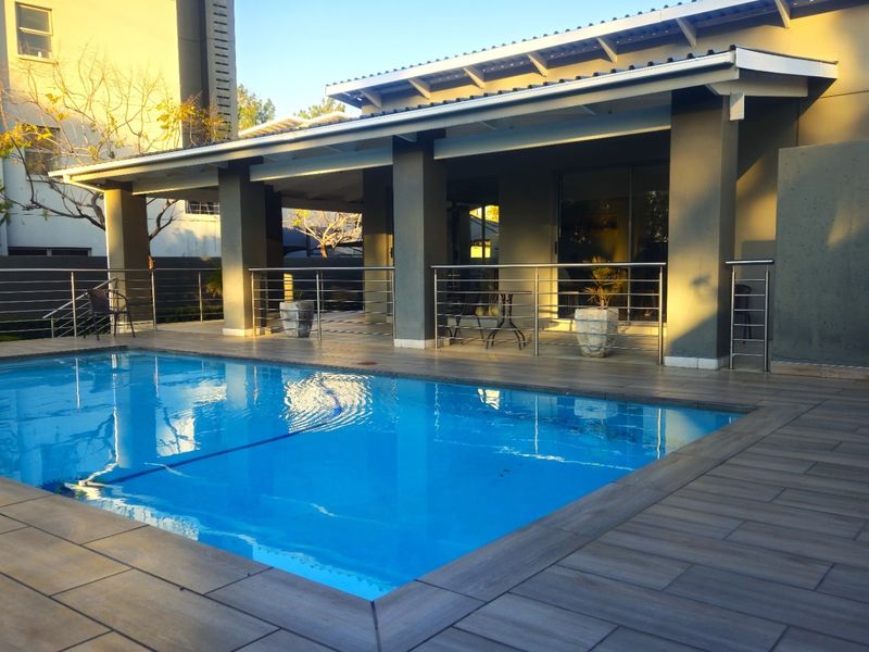 A well-maintained outdoor pool area featuring a tiled deck, covered patio space, and modern stainless steel balustrades.