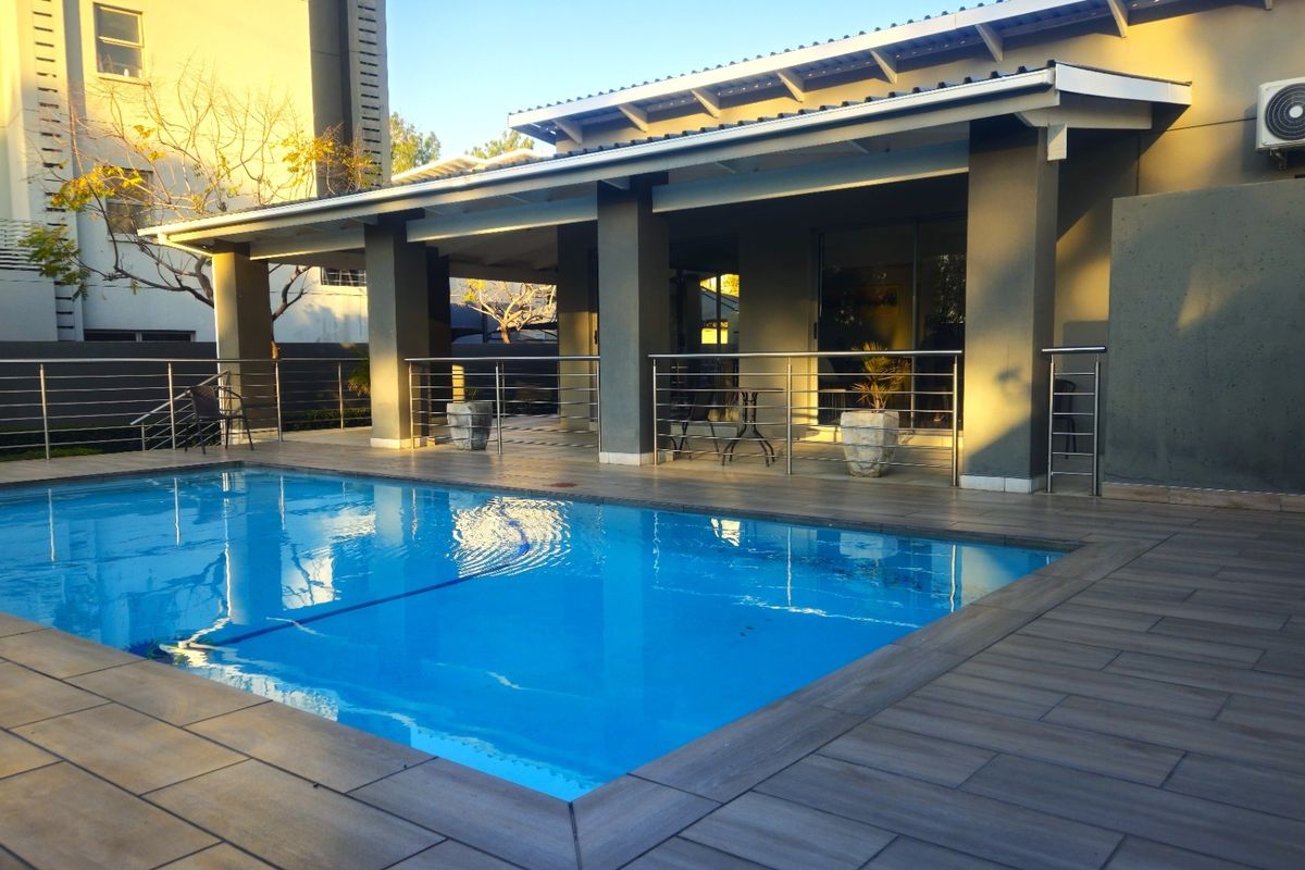 A well-maintained outdoor pool area featuring a tiled deck, covered patio space, and modern stainless steel balustrades.