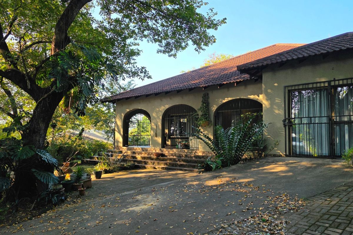Rear exterior view of the house featuring a tiled roof, arched patio openings, security gates, and a paved outdoor area surrounded by mature trees.