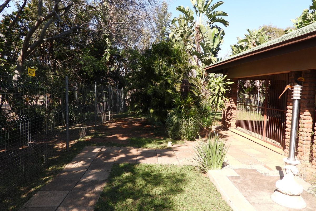 A rear garden area featuring a paved patio, brick exterior wall, and secure wire mesh fencing surrounded by established trees and landscaping.