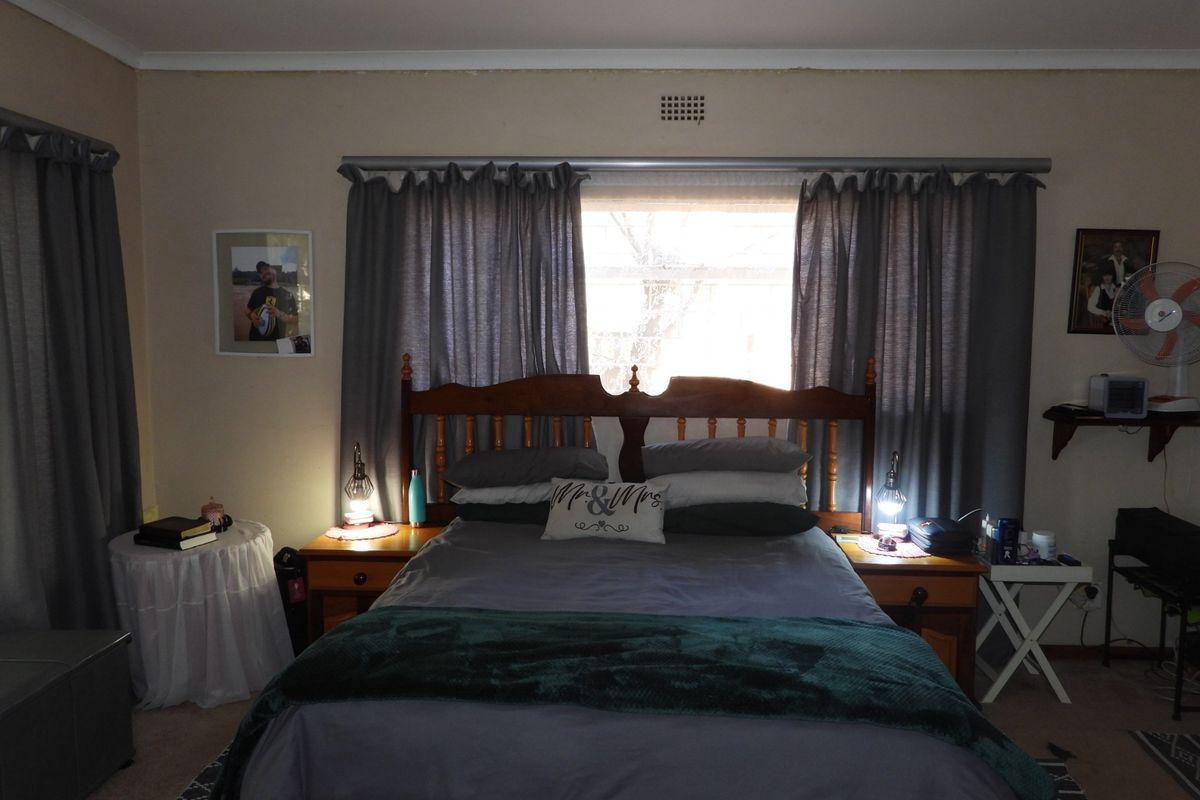 A standard bedroom featuring a central window with curtain rod and wall-mounted air vent, showing signs of wear near the ceiling cornice.