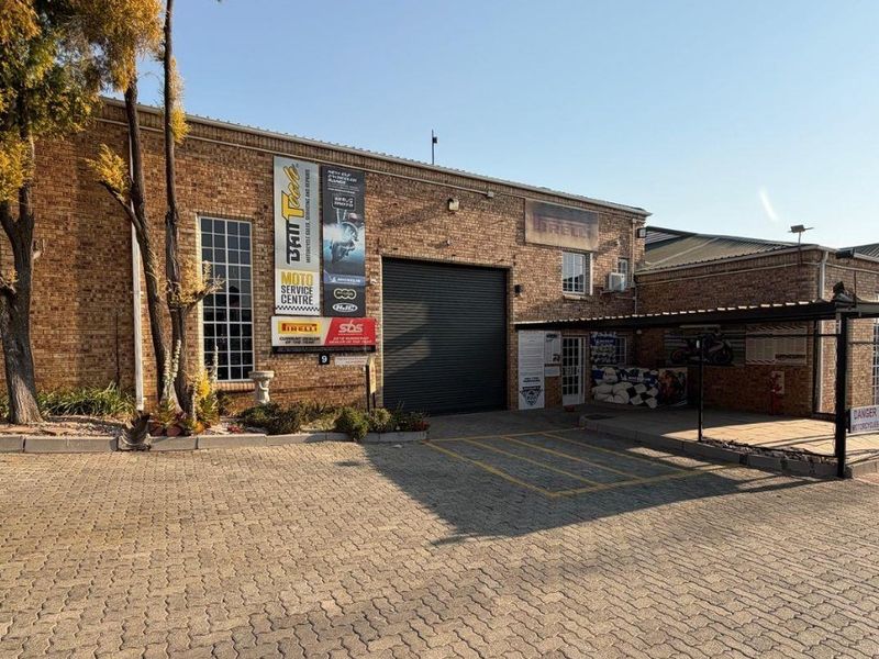 Front exterior view of an industrial property featuring a brick facade, roller shutter door, and a paved driveway with covered parking.