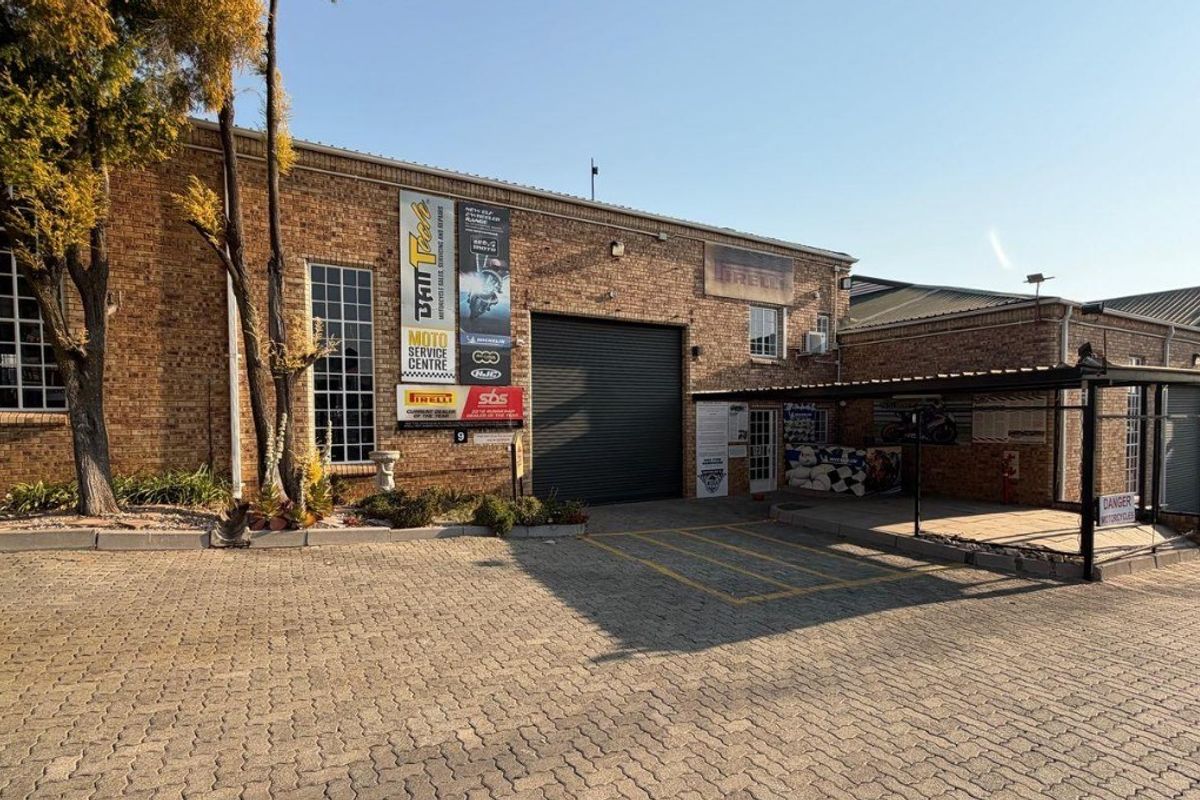 Front exterior view of an industrial property featuring a brick facade, roller shutter door, and a paved driveway with covered parking.