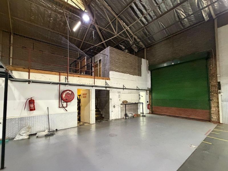 Spacious industrial utility area featuring a large roller shutter door, mezzanine level, exposed roof trusses, and durable concrete flooring suitable for workshop or storage use.