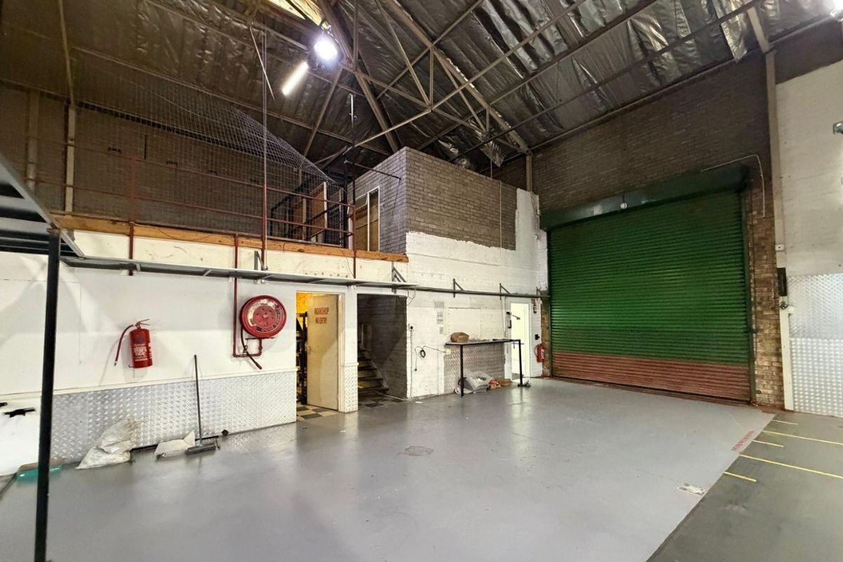 Spacious industrial utility area featuring a large roller shutter door, mezzanine level, exposed roof trusses, and durable concrete flooring suitable for workshop or storage use.