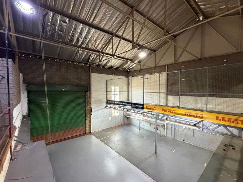 Spacious industrial warehouse area featuring high ceilings with exposed steel trusses, a roller shutter door, and a functional mezzanine level with safety railing.
