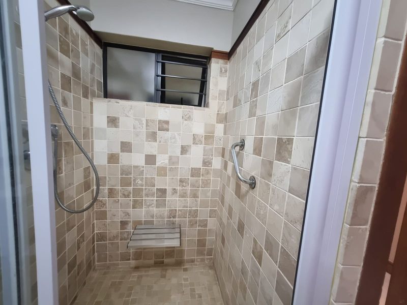 A clean, tiled shower enclosure featuring a built-in seat, safety grab rail, and a louvered window for ventilation.