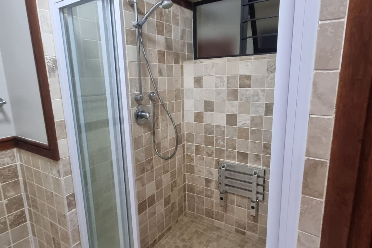 A functional bathroom shower area featuring neutral-toned wall and floor tiles, a built-in shower seat, and a louvered window for ventilation.